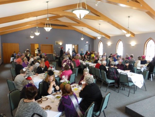 Fascinator tea party – 50 people in fancy hats seated at tables enjoying tea and sandwiches.